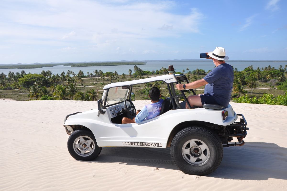 Pessoas andando de buggy vermelho numa trilha de areia, perto do mangue.
